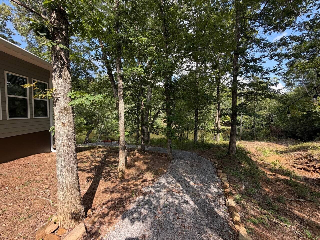 Un chemin de gravier traverse une zone boisée, avec des arbres et un bâtiment situé sur la gauche.
