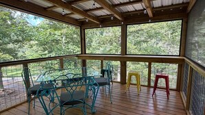 Un espacio al aire libre techado con una terraza de madera, sillas de estructura metálica y una mesa.