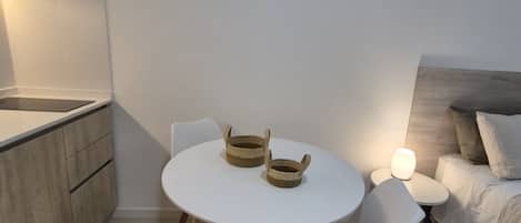 A small, modern room with a round white table, two chairs, a bedside table with a lamp, a bed with a headboard, and a wooden cabinet.