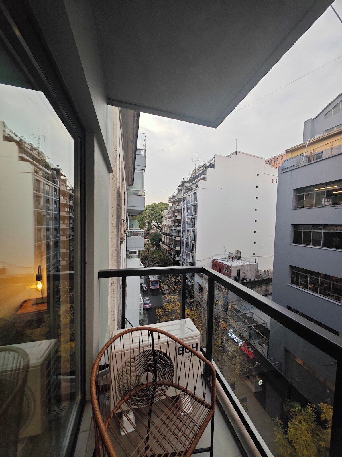 Ein Balkon mit einem Korbstuhl, einem Ventilator und Blick auf eine Stadtlandschaft mit Gebäuden.