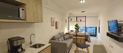 A modern kitchen with a built-in microwave, a coffee machine, and a sink. There is a grey sofa and a glass table with chairs in the living area.