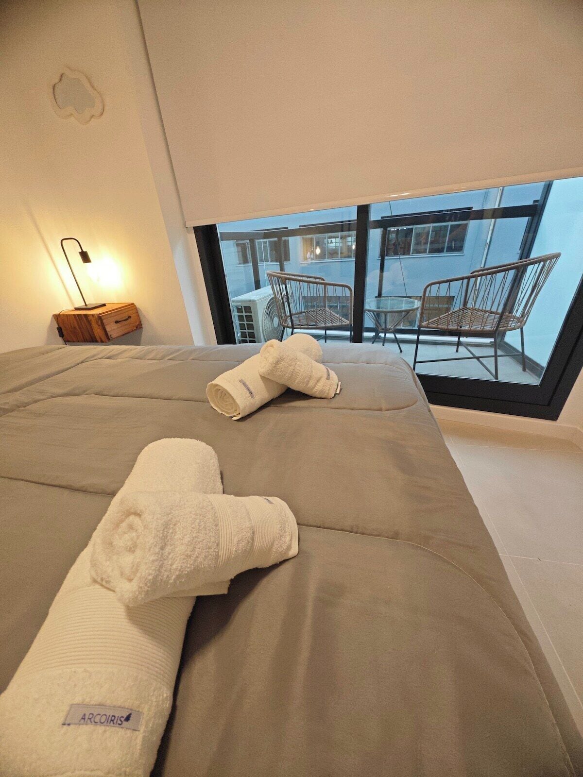 A bed with two rolled towels, a bedside lamp, and a view of an outdoor seating area through a large window.