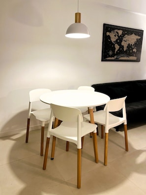 Ein Essbereich mit einem runden weißen Tisch und vier Stühlen mit Holzbeinen. Eine schwarze Couch steht an der Wand, darüber ist eine Weltkarte angebracht.