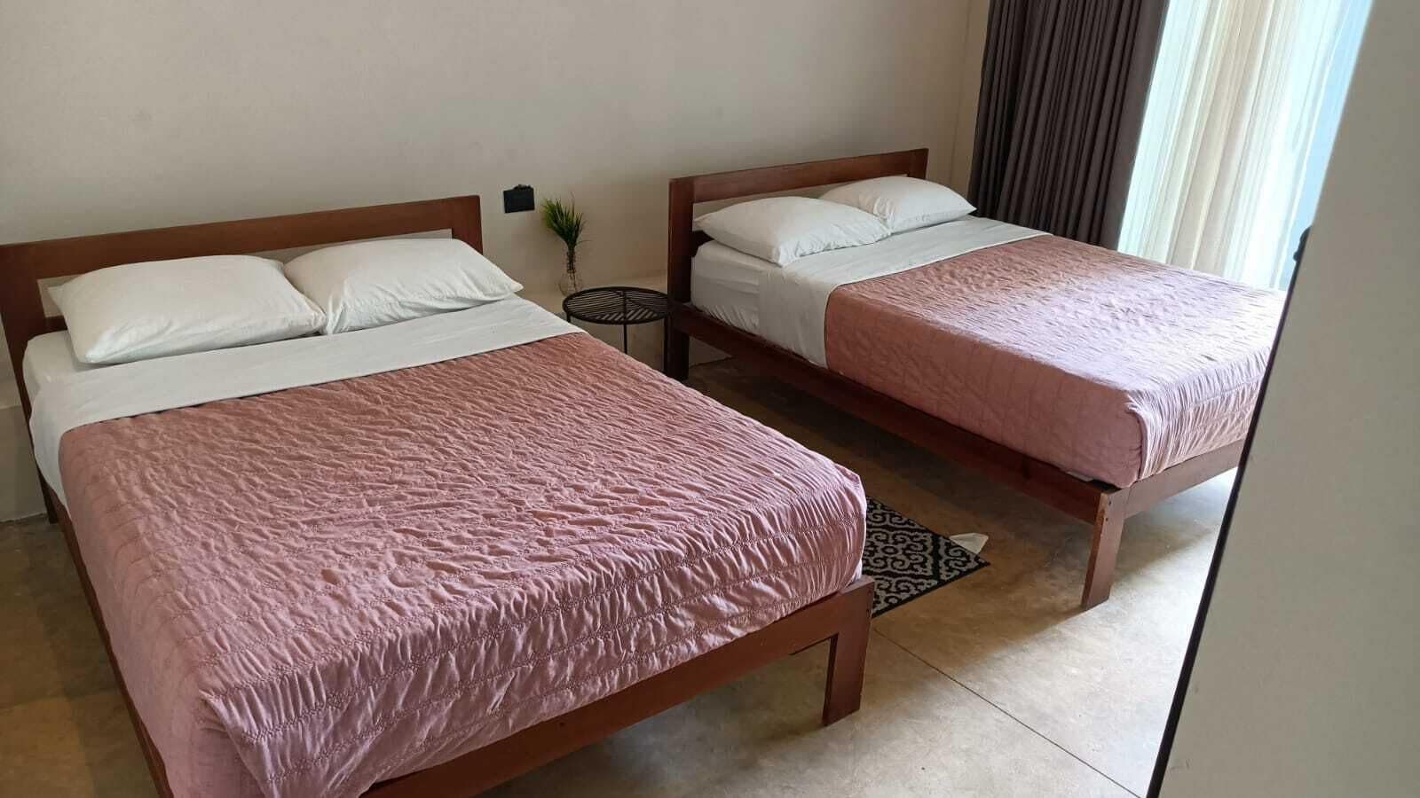 Dos camas individuales con cabeceros de madera y ropa de cama blanca, una mesita redonda pequeña y una alfombra estampada sobre suelo de baldosas.