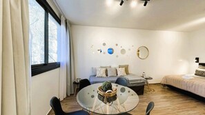 Un dormitori modern amb llit, taules de nit, una taula rodona de vidre amb cadires i una finestra amb cortines.