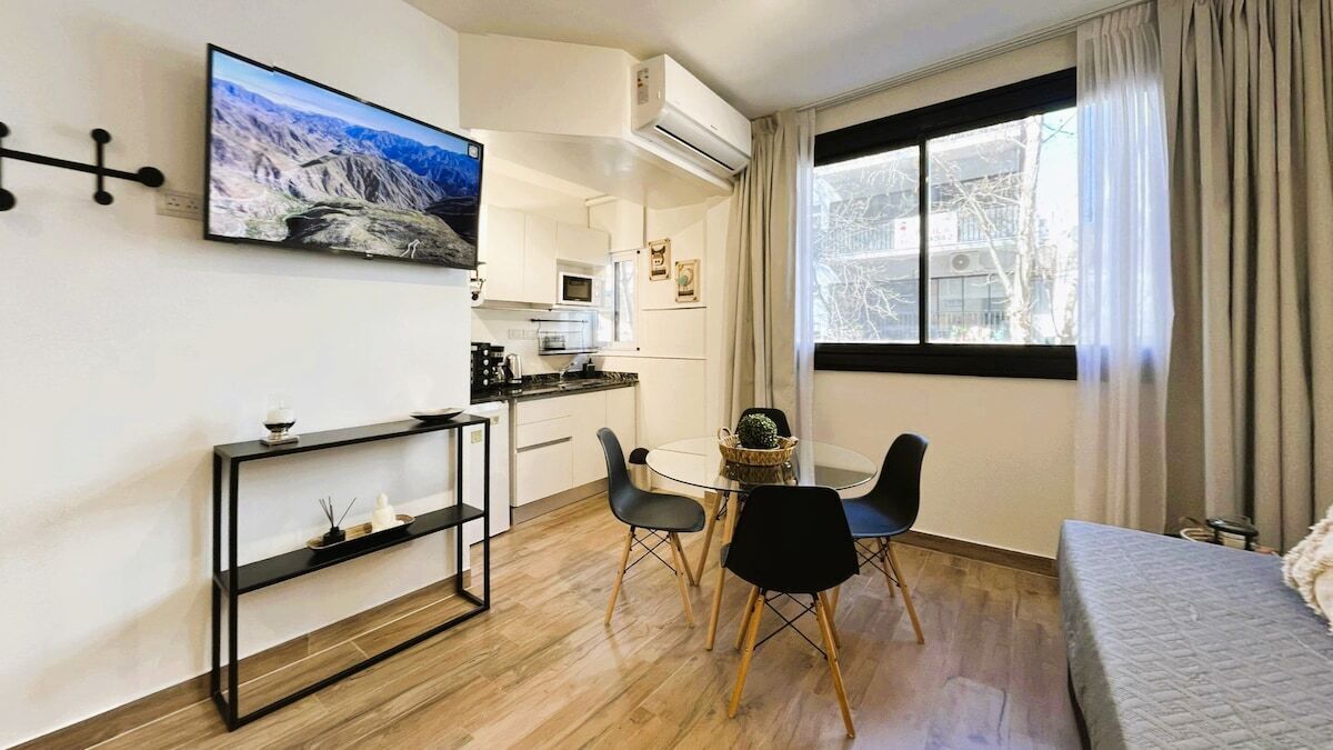 A compact living space with a kitchenette, a dining area with a table and chairs, a bed, and a large window with curtains.