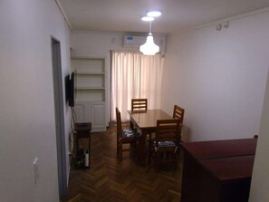 Uma sala de jantar com mesa e cadeiras de madeira, luminária no teto e janela com cortinas.
