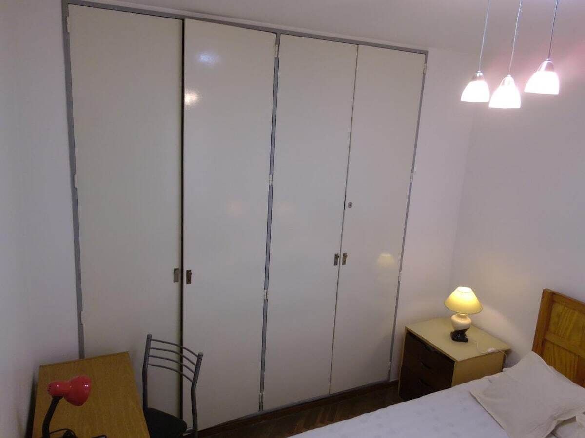 A bedroom with a wardrobe, a bedside table with a lamp, a chair, and a bed.