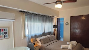 Quarto com cama, mesa de cabeceira, abajur, janela com cortinas, porta e ar-condicionado fixado na parede.