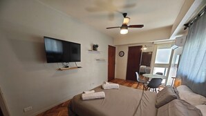 Quarto com cama, televisão na parede, ventoinha de teto e pequena mesa com cadeiras.