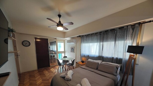 Quarto de hotel com cama, ventoinha no teto, escrivaninha com cadeira e janela com cortinas.