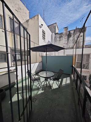 Une petite terrasse extérieure avec une table ronde et des chaises, un parasol noir et une clôture en métal.