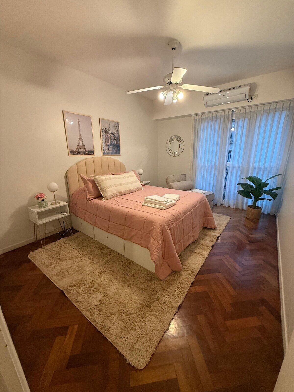 A bedroom with a bed, a bedside table, a nightstand, a ceiling fan, a wall clock, and a window with curtains.