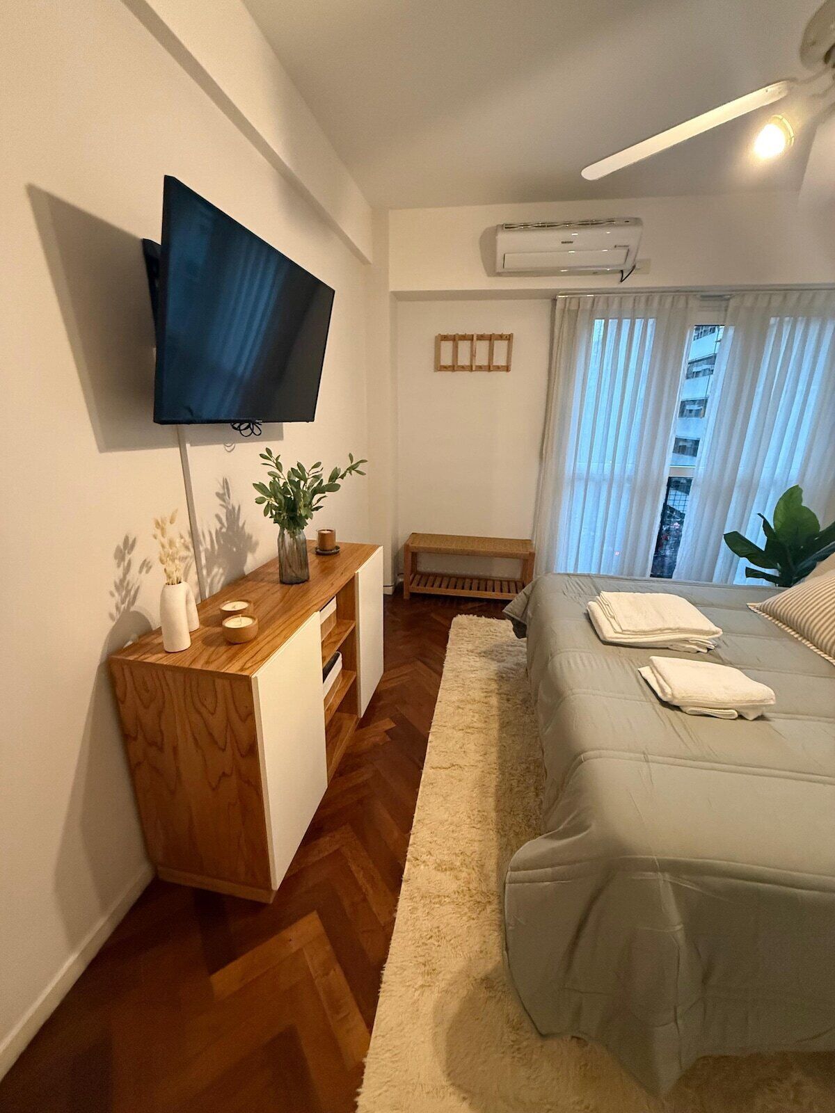 Une chambre moderne avec un téléviseur à écran plat fixé au mur, un bureau en bois agrémenté d’une plante, un lit avec des serviettes soigneusement pliées et une fenêtre ornée de rideaux légers.