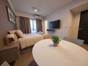 Ein modernes Hotelzimmer mit Bett, Nachttisch, Lampe, Fernseher und einem runden Tisch mit Pflanze.