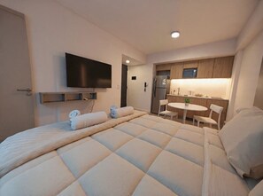 Habitación de hotel con una cama grande, un televisor de pantalla plana en la pared, una kitchenette con refrigerador y una pequeña mesa de comedor con sillas.