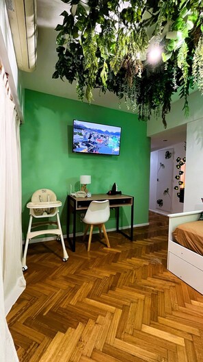 A room with a green wall, a mounted television, a desk with a chair, a white baby high chair, and a bed with a brown mattress. - Jungle Chic in Palermo · Vibrant & Unique (Buenos Aires)