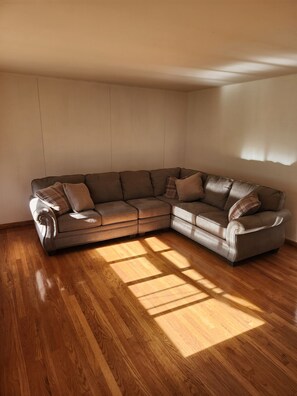 A corner living room with a brown leather sectional sofa, wooden flooring, and a plain wall.