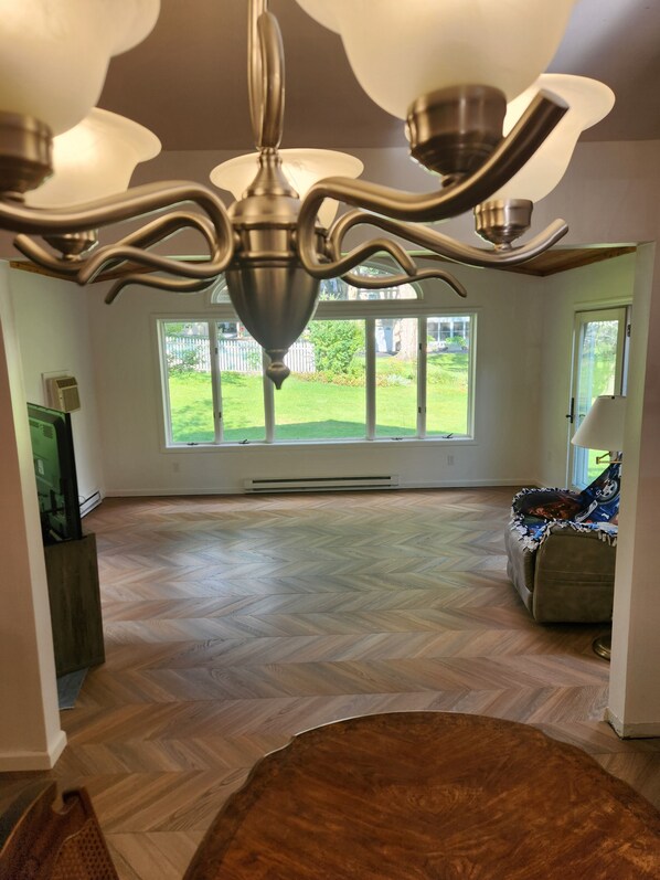 A spacious room with a herringbone floor, a ceiling fan, and a large window offering a view of a green yard.