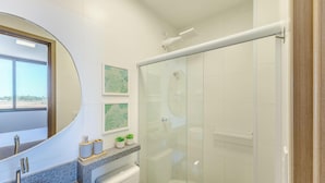 A modern bathroom with a glass shower enclosure, a large mirror, and a window providing a view of the outdoors. - Apto Encantador Cerca del mar en Marau Vll0107 (Maraú)