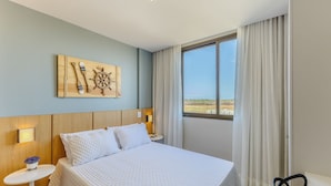 A hotel room with a bed, a wall-mounted artwork, a window with curtains, a bedside table with a lamp, and a small potted plant. - Apto Encantador Cerca del mar en Marau Vll0107 (Maraú)