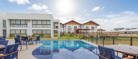 Una piscina con mosaicos azules, rodeada de sillones y mesas, con edificios al fondo.