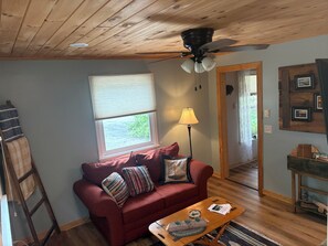 A cozy living room with a wooden ceiling, a red sofa with cushions, a wooden coffee table, a floor lamp, and a window with blinds.