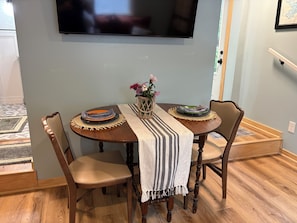 A dining area with a round table, two chairs, a TV mounted on the wall, and a framed map on the wall. - Cozy 1-bedroom vacation cottage on beautiful Lake Ann (Lake Ann)
