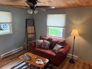 A living room with a red sofa, a wooden coffee table, a floor lamp, and a ceiling fan. - Cozy 1-bedroom vacation cottage on beautiful Lake Ann (Lake Ann)