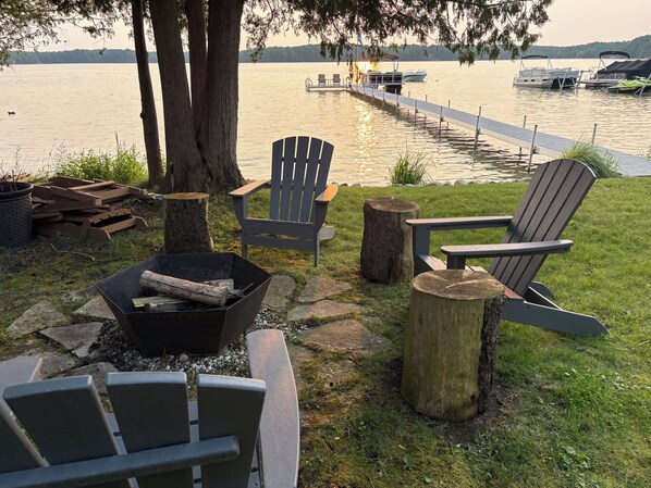 A lakeside deck with Adirondack chairs, a fire pit, and a dock extending into the water.
