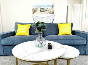 Ein Wohnzimmer mit einem blauen Sofa, zwei gelben Kissen, einer Glasvase mit Grünpflanzen und einer Fernbedienung auf einem weißen Marmor-Couchtisch.