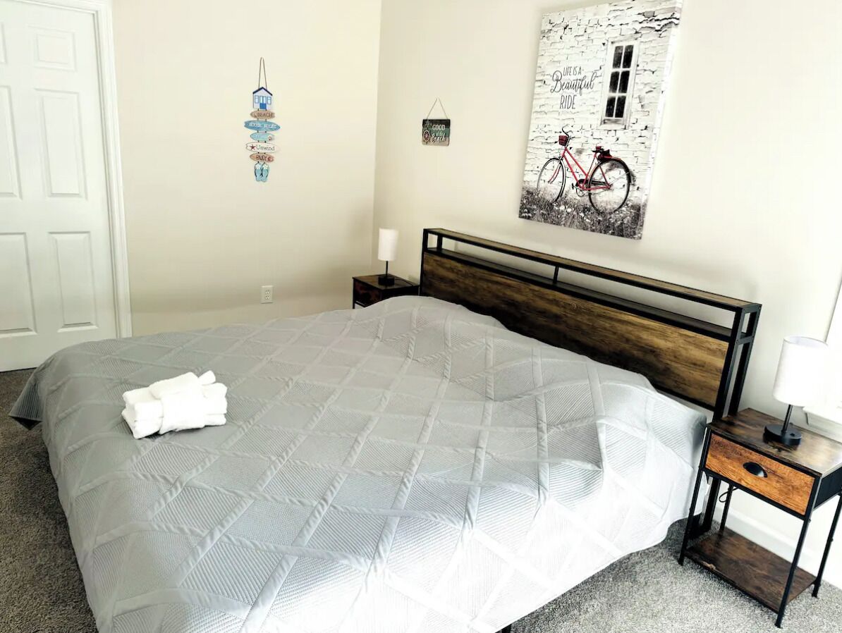 Uma cama bem arrumada com um edredom cinza, uma cabeceira de madeira, uma mesa de cabeceira com abajur e uma parede com arte decorada com tema de bicicleta.