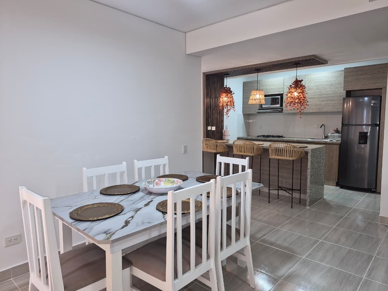 Una cocina moderna con comedor, que incluye una mesa de mármol, sillas blancas y un refrigerador de acero inoxidable.