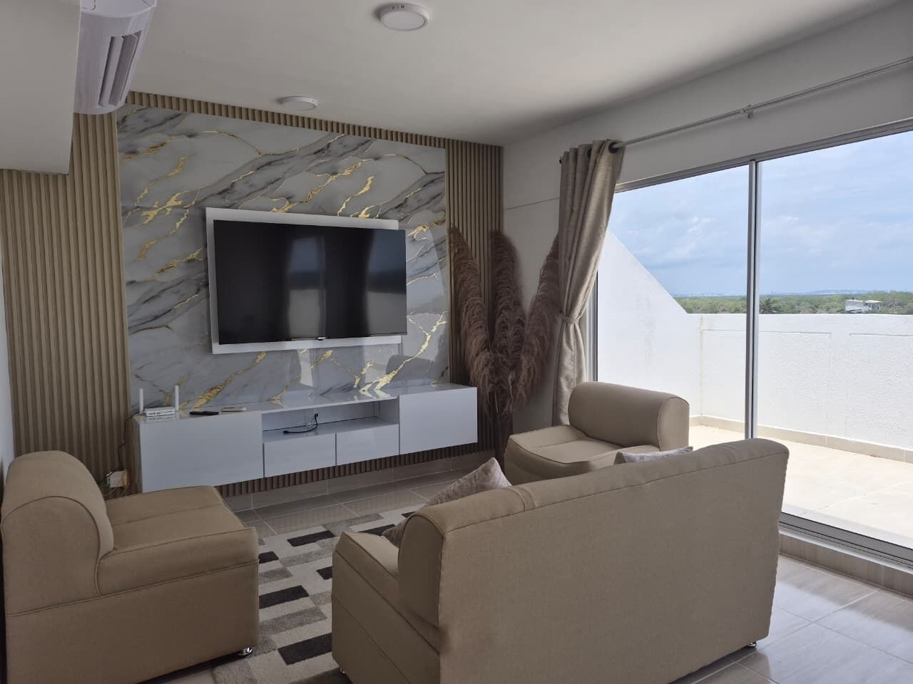 A modern living room with a flat-screen TV mounted on a marble wall, a sofa set, and a large window offering a view.