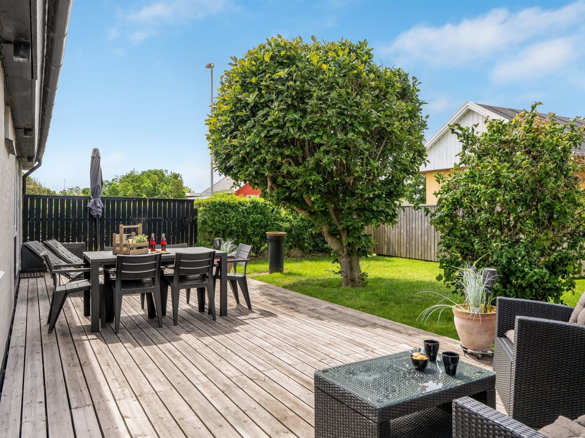 A wooden deck with a table and chairs, surrounded by greenery and a fenced yard.