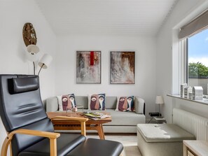 Ein modernes Wohnzimmer mit Ledersessel, Couchtisch aus Holz, grauem Sofa mit gemusterten Kissen und abstrakten Bildern an der Wand.
