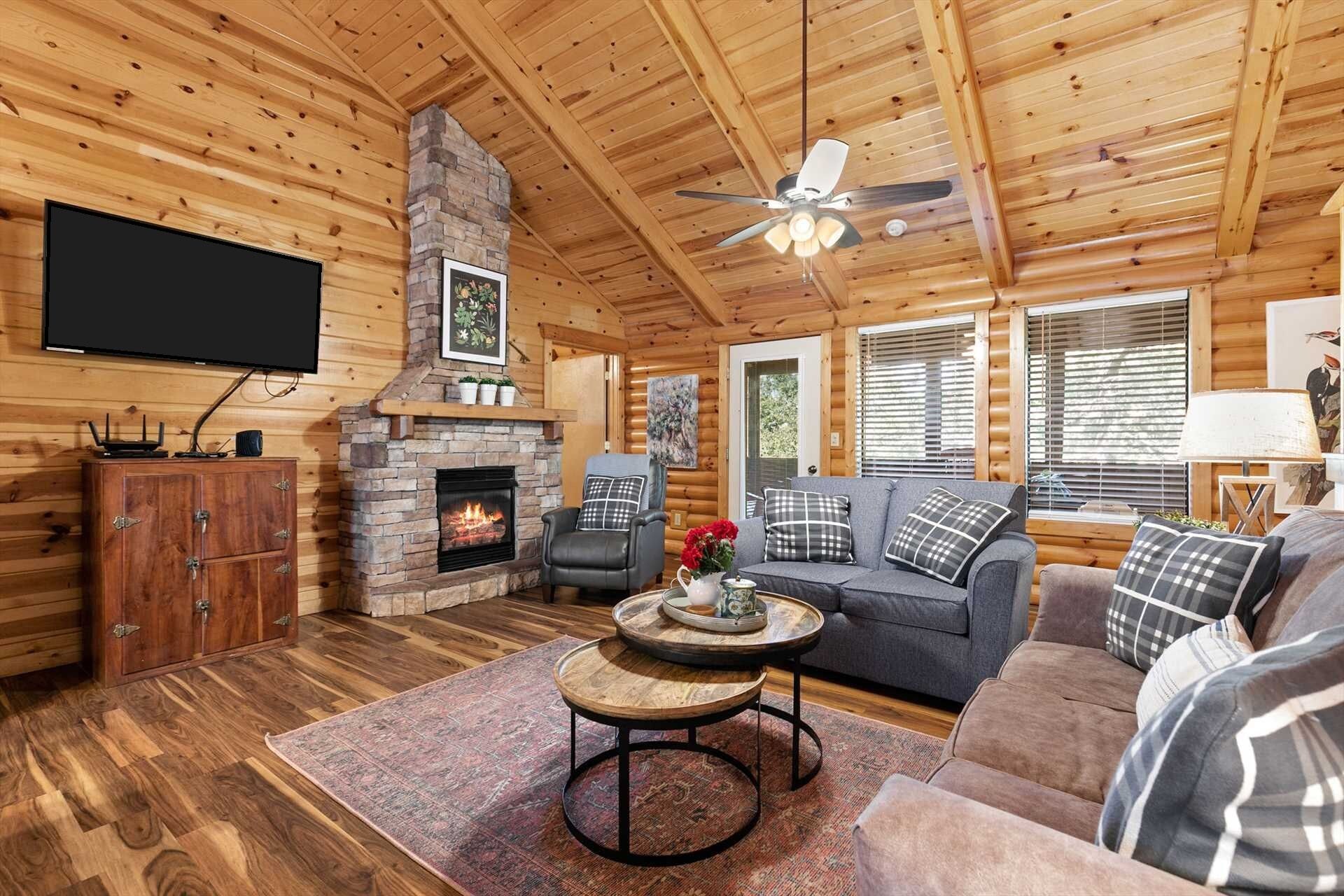 A cozy cabin interior with a stone fireplace, a flat-screen TV, a sofa, and a ceiling fan.