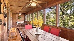 A wooden interior with a dining table, red chairs, a ceiling fan, and large windows offering a view of trees.