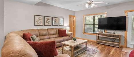 A living room with a leather sofa, a coffee table, a TV on a wooden stand, and framed botanical prints on the wall.