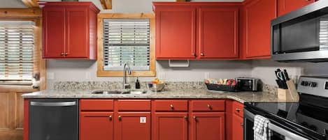 Una cocina con gabinetes rojos, encimera de granito, pileta de acero inoxidable, microondas y horno.