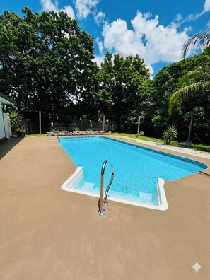 Una piscina rectangular amb vora blanca, envoltada d'un sòcol de formigó i amb arbres al fons.