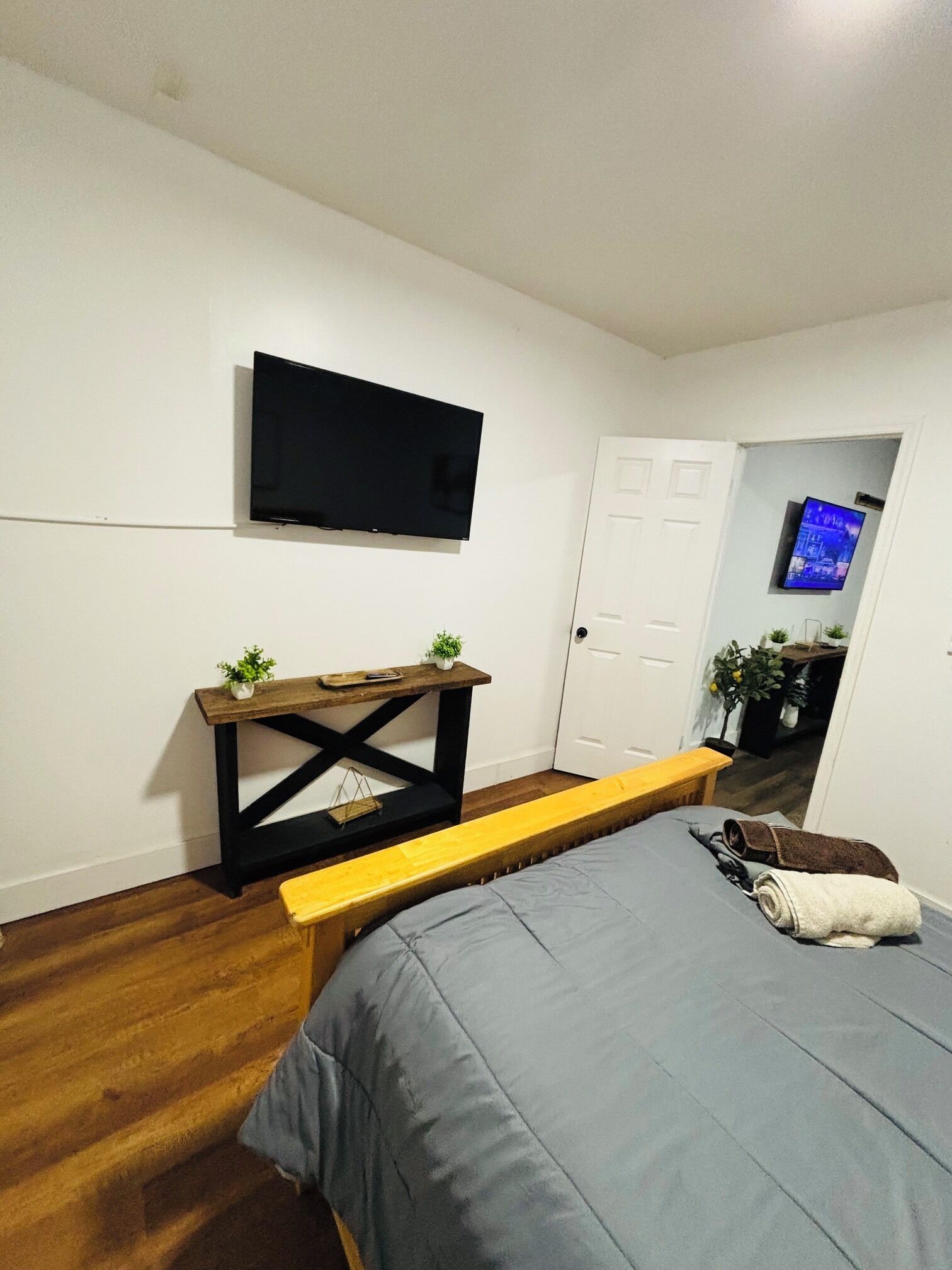 Une chambre à coucher comprenant un lit, un banc en bois, un téléviseur fixé au mur et une porte donnant sur une autre pièce.