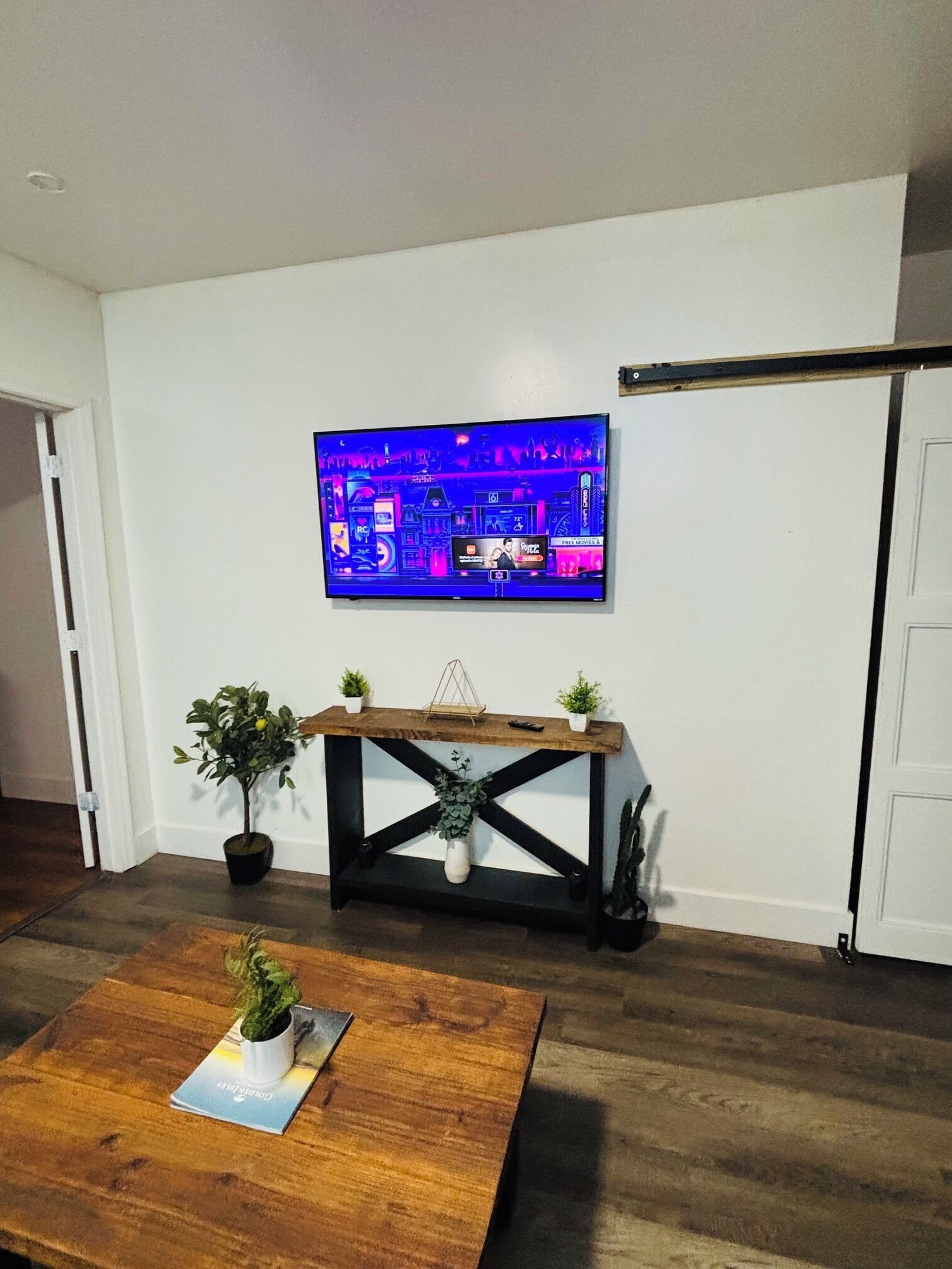 Una sala de estar con un televisor de pantalla plana montado en la pared, una mesa de centro de madera con una revista y una pequeña planta en un lateral.