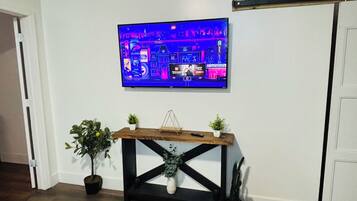Una sala de estar con un televisor de pantalla plana montado en la pared, una mesa de centro de madera con una revista y una pequeña planta en un lateral.