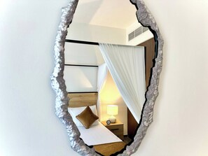 Een spiegel met een bijzondere, onregelmatige lijst die een kamer weerkaatst met een bed, nachtkastje, lamp en gordijnen.