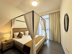 Ein Hotelzimmer mit Himmelbett, Nachttisch mit Lampe und Blick auf die Stadt durch große Fenster.