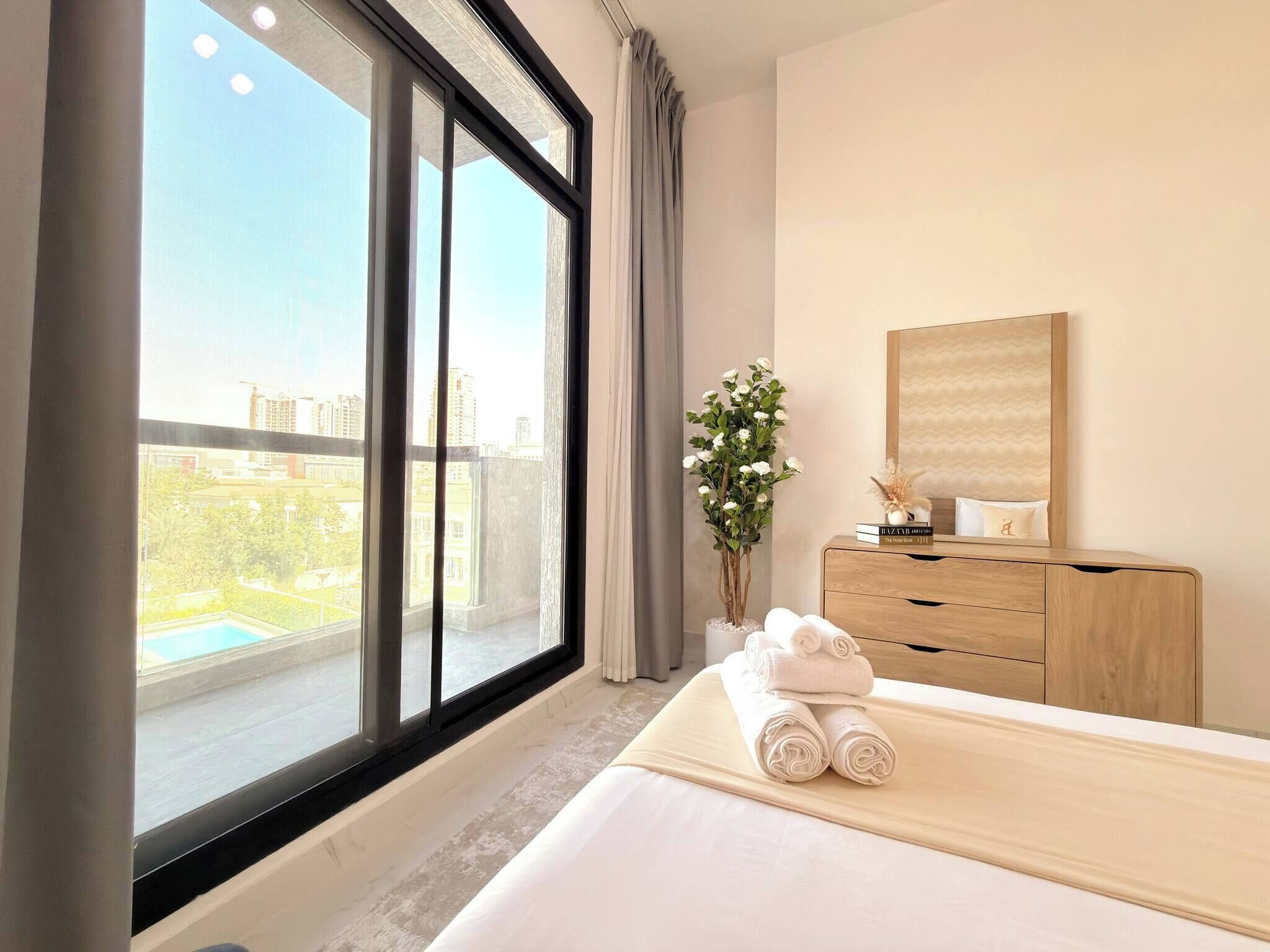 A hotel room with a large window, a bed with rolled towels, a wooden dresser, a vase with flowers, and a mirror on the wall.