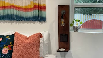 Un dormitorio con cama, almohadas, un tapiz en la pared, una lámpara de pared, un enchufe, una ventana con vistas a la vegetación y una planta en el alféizar.
