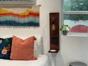 A bedroom with a bed, pillows, a wall hanging, a wall-mounted light, a power outlet, a window with a view of greenery, and a potted plant on the windowsill.