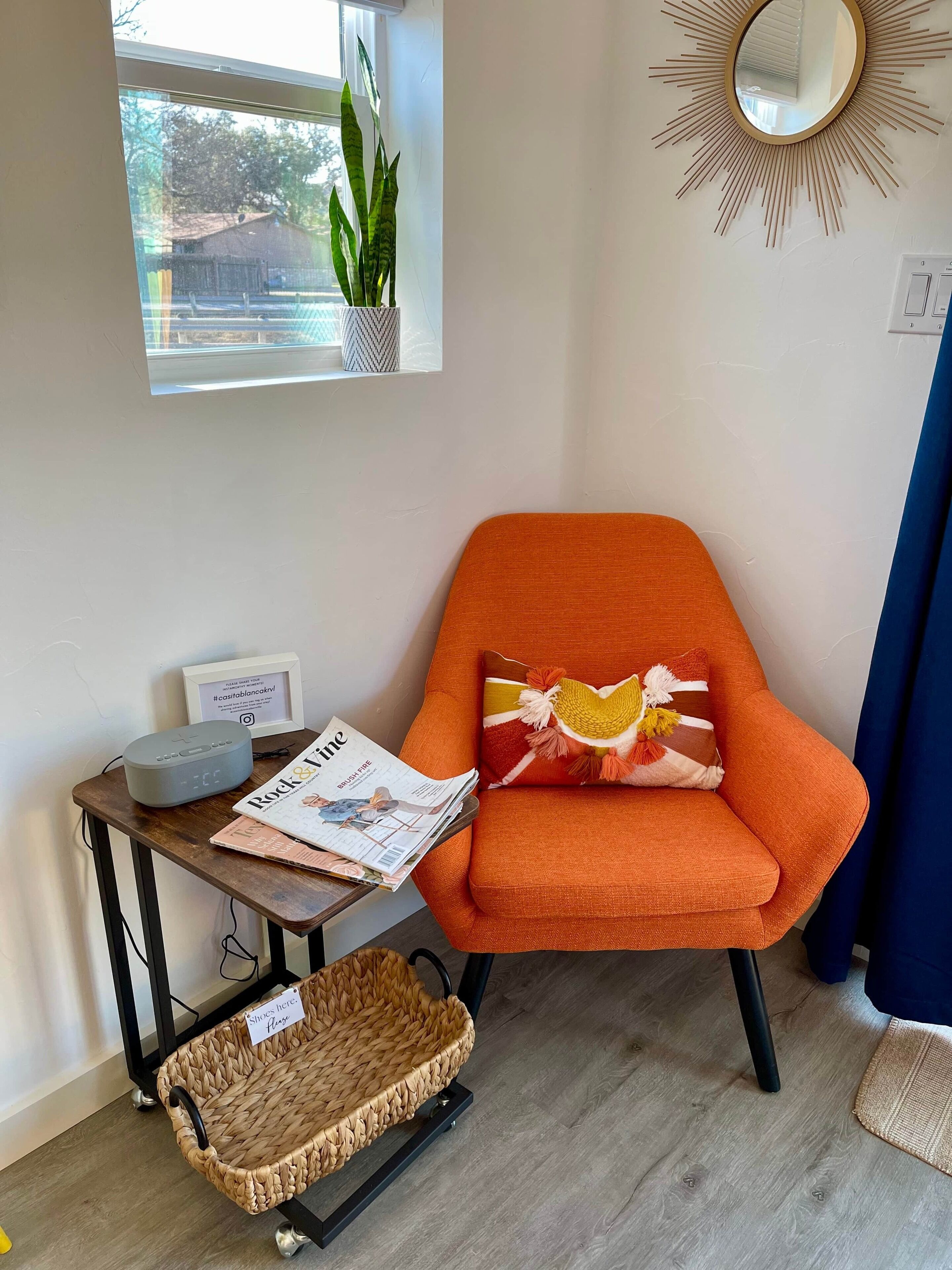 A corner room with a window, a plant, a mirror, a chair, a table with a magazine, a speaker, and a basket.